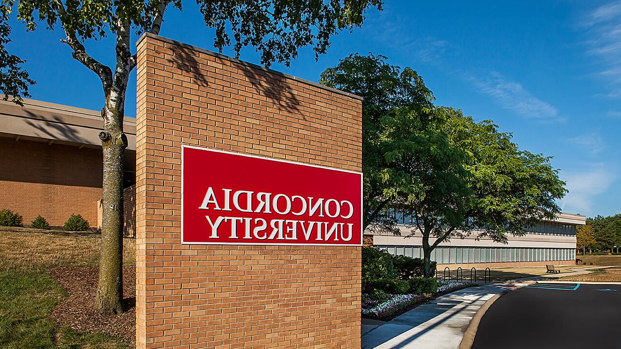 A sidewalk that runs along the parking lot leads to the main campus entrance; surrounded on one side by a small flower garden and a few trees, and on the other side by a wall which extends from the front entrance; greeting visitors with a large bold Cardinal-red Concordia University sign.